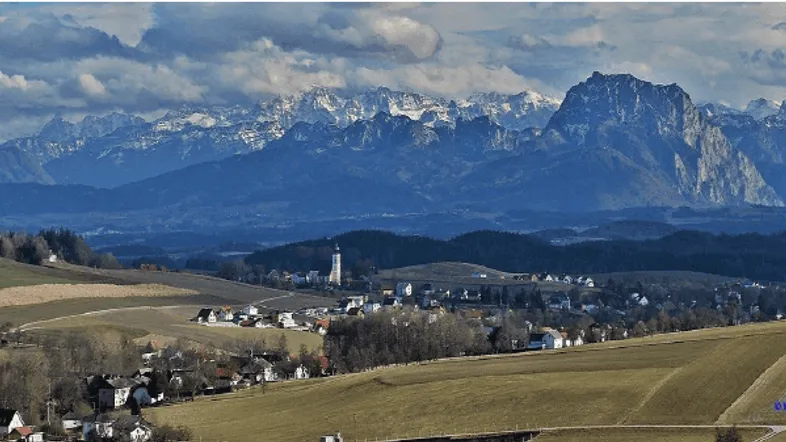 Panoramablick auf ein Dorf in einem Tal mit schneebedeckten Bergen im Hintergrund und weiten Feldern.