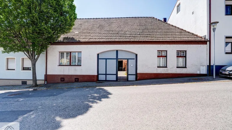 Vorderansicht eines traditionellen Hauses mit weißer Fassade und braunem Dach in ländlicher Umgebung.