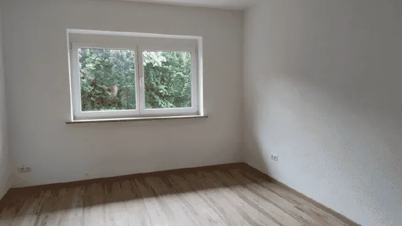 Helles, leeres Zimmer mit Holzboden und Fensterblick ins Grüne, bereit zur individuellen Gestaltung.