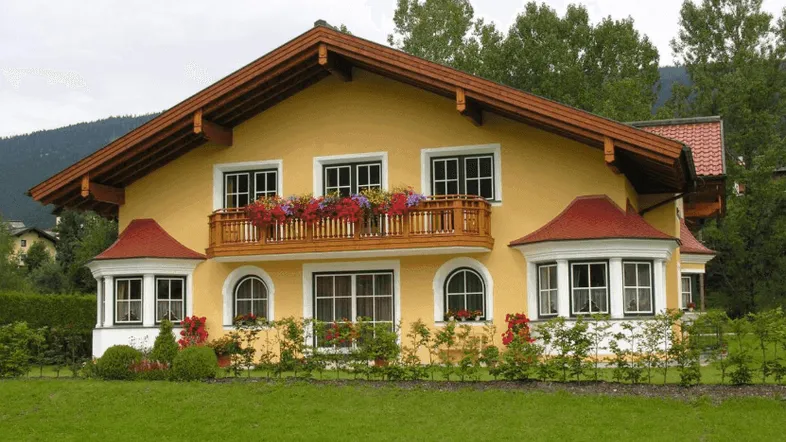 Gelbes Haus mit traditioneller Holzfassade, Balkon mit Blumenkästen und grüner Rasenfläche.