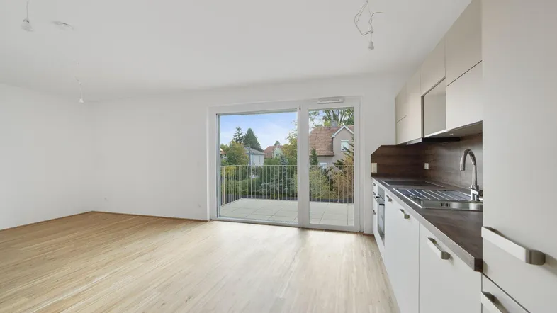 Helles Zimmer mit Holzboden, integrierter Küche und Zugang zu einem Balkon mit Blick ins Grüne.