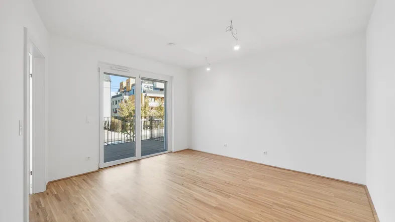 Helles, leeres Zimmer mit Holzboden und Zugang zu einem Balkon, ideal als Wohn- oder Schlafzimmer.