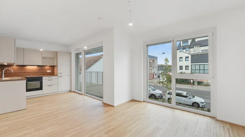 Heller offener Wohnbereich mit integrierter Küche, Parkettboden und Zugang zum Balkon.