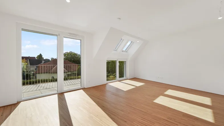 Helles Dachgeschosszimmer mit Holzboden, großen Fenstern und Zugang zu einem Balkon, lichtdurchflutet von Sonnenstrahlen.