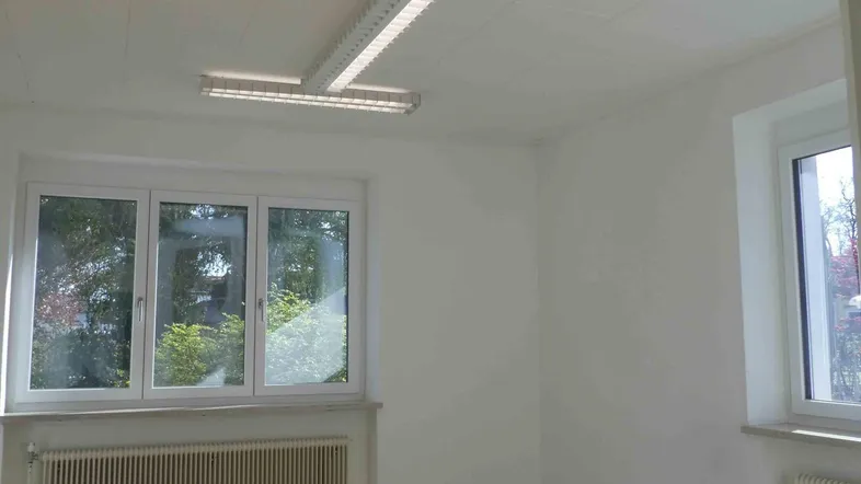 Heller Büroraum mit großen Fenstern, Laminatboden und effizienten Deckenleuchten, ideal für produktives Arbeiten.