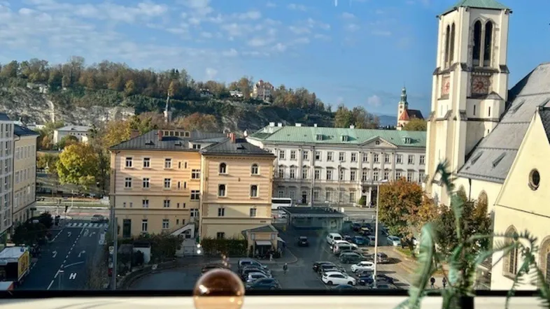 Panoramablick aus einem Fenster auf die historische Stadt Salzburg mit Bergen im Hintergrund.