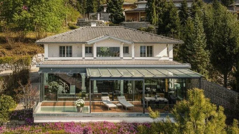 Haus mit moderner Glasfassade, umgeben von blühenden Gärten und Bergen im Hintergrund.