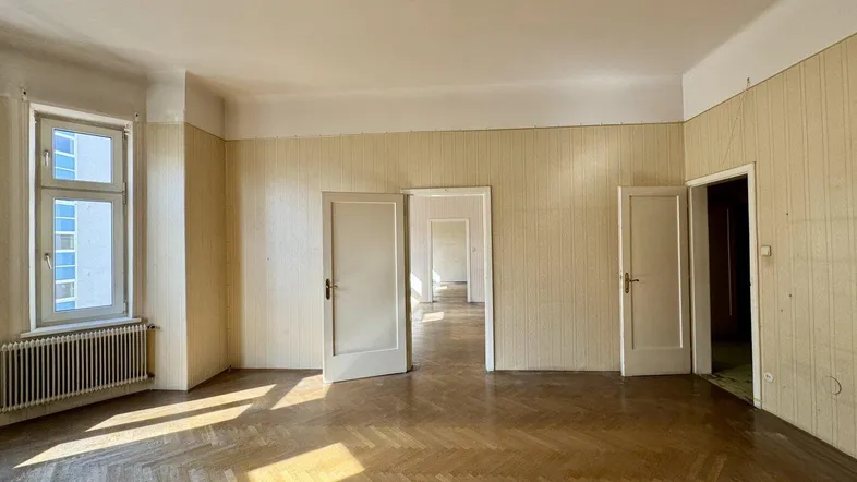 Helles Wohnzimmer mit Fischgrätparkettboden, hohen Decken und Tageslicht durch ein großes Fenster.