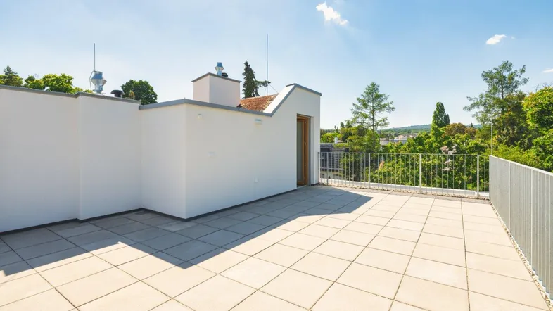 Große Dachterrasse mit hellen Fliesen, weißer Brüstung und weitem Blick ins Grüne unter blauem Himmel.