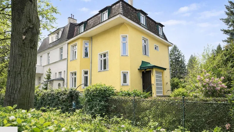 Gelbe Villa mit Mansarddach und umgebendem Garten in grüner Umgebung, umgeben von Bäumen.