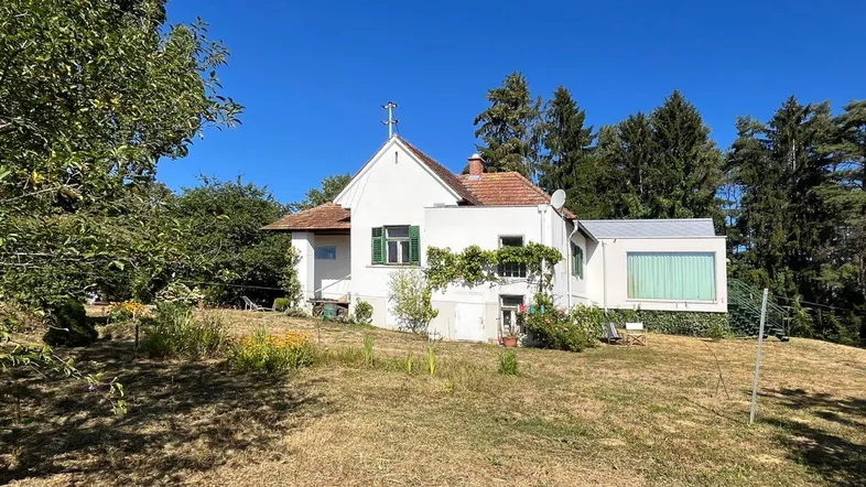 Weißes Haus mit rotem Ziegeldach, grünen Fensterläden und modernem Anbau in ländlicher Umgebung.