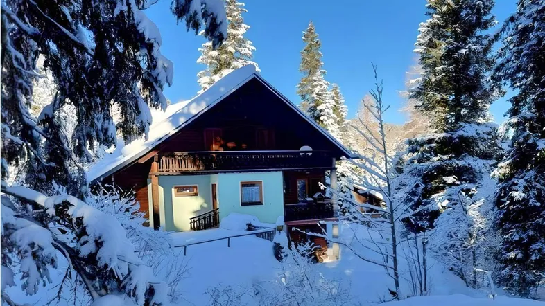 Charmantes Chalet in verschneiter Winterlandschaft, umgeben von hohen Tannenbäumen unter blauem Himmel.