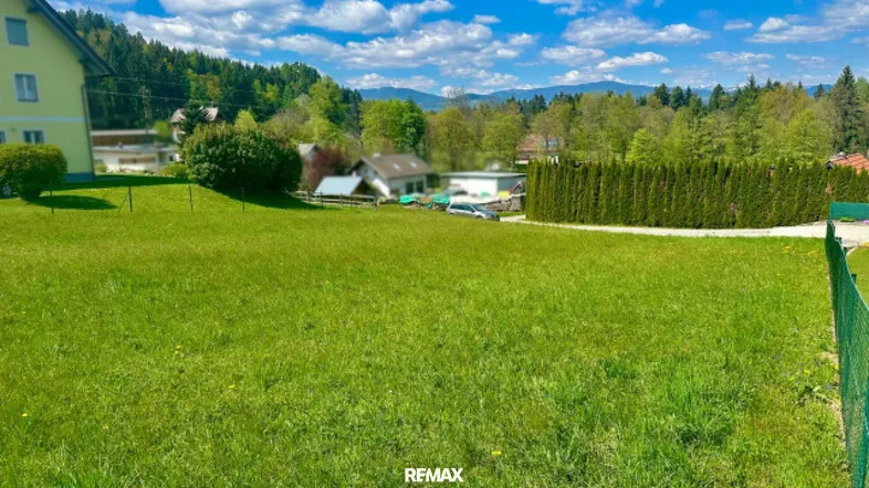 Weitläufige grüne Wiese mit Blick auf umliegende Häuser und bewaldete Hügel unter blauem Himmel.