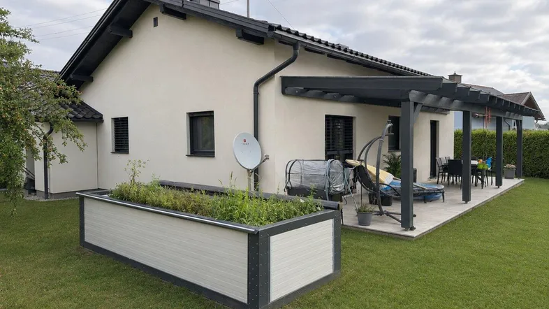 Hausfassade mit gepflegtem Rasen, Hochbeet und überdachter Terrasse mit Sitzgelegenheiten im Freien.
