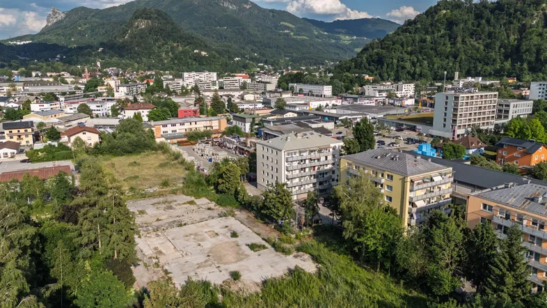 Luftaufnahme einer Stadtlandschaft mit Wohngebäuden, viel Grün und Bergen im Hintergrund, inklusive einer großen Freifläche.