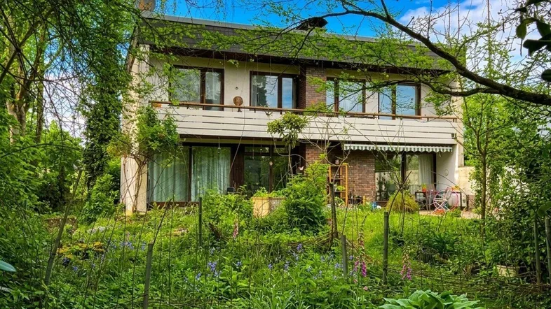 Hausfassade umgeben von üppigem Grün und Bäumen mit sichtbaren Balkonen.