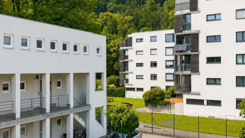 Moderne Wohngebäude mit weißer Fassade, umgeben von grünen Hügeln und einem Spielplatz.