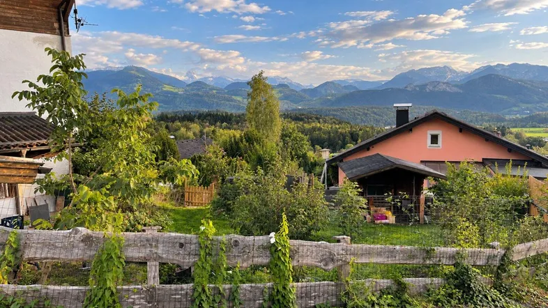 Blick auf ein Haus mit Garten, umgeben von grüner Landschaft und majestätischen Bergen im Hintergrund.