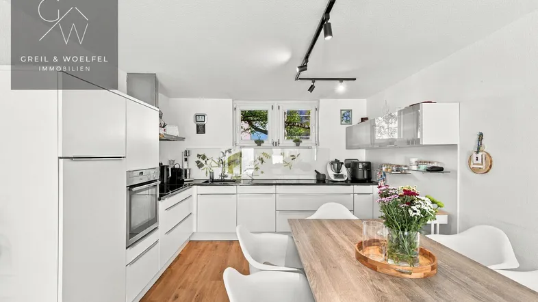 Moderne Küche mit weißen Schränken, Holztisch und Blick ins Grüne durch das Fenster.