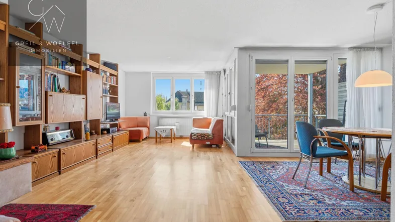 Geräumiger Wohn-Essbereich mit Parkettboden, großem Bücherregal und Zugang zum Balkon.