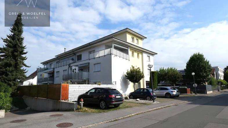 Mehrfamilienhaus mit mehreren Balkonen und Parkplätzen vor dem Gebäude unter blauem Himmel.