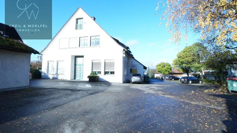 Weißes Gebäude mit Satteldach und großem gepflasterten Vorplatz, umgeben von Bäumen mit Herbstlaub und parkenden Autos.