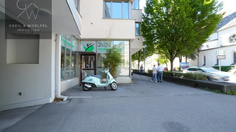 Außenansicht eines Geschäftsgebäudes mit Schaufenstern und einem Roller vor dem Eingangsbereich.