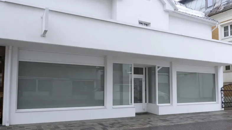 Modernes weißes Geschäftsgebäude mit großen Schaufenstern, Rollläden und einem gepflegten Eingangsbereich.