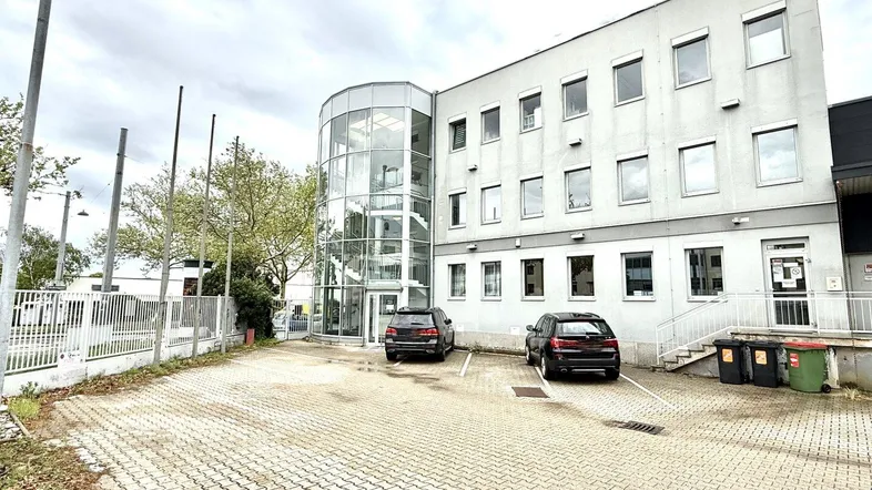 Modernes Bürogebäude mit Glasfassade und Parkplätzen davor unter bewölktem Himmel.