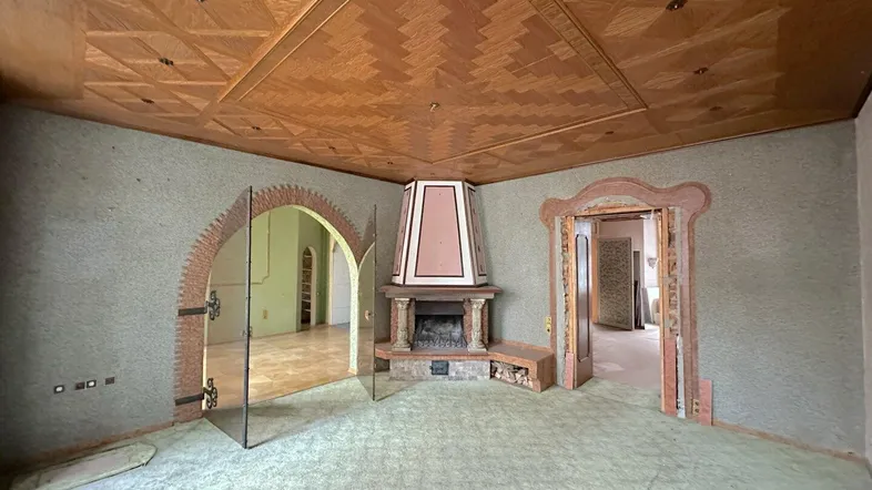 Geräumiges Wohnzimmer mit gemusterter Holzdecke, Kamin und Teppichboden, bereit zur Renovierung.