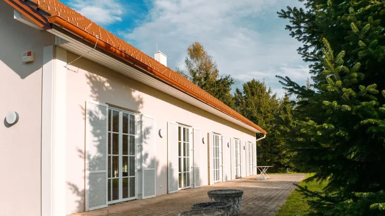 Helle Hausfassade mit mehreren Fenstertüren, die sich zu einer gepflasterten Terrasse öffnen.