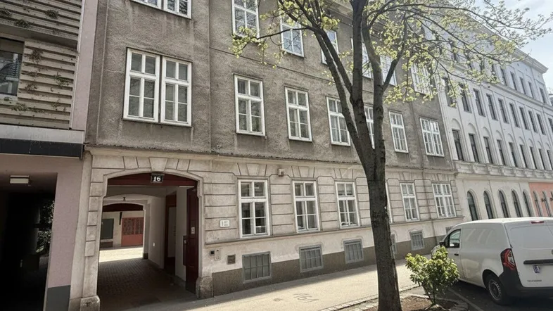 Graue Fassade eines Altbaus mit vielen Fenstern und einem Durchgang zum Innenhof, umgeben von Bäumen.