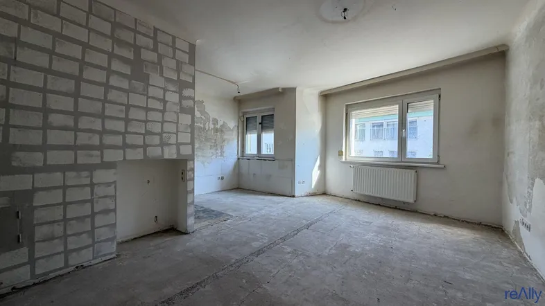 Unrenovierter Wohnraum mit freigelegten Wänden, Betonboden und Heizkörper unter den Fenstern.