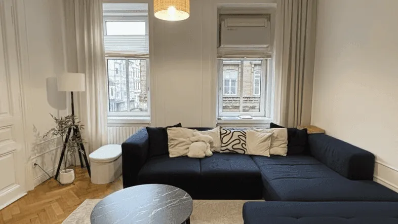 Gemütliches Wohnzimmer mit dunkelblauem Ecksofa, Parkettboden und zwei Fenstern zur Straße.