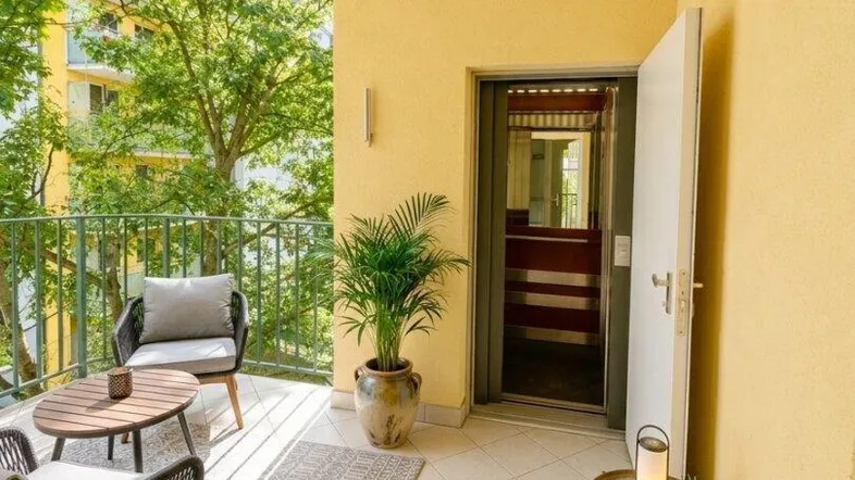 Möblierter Balkon mit gelber Fassade, Pflanzen und direktem Zugang zu einem Aufzug.