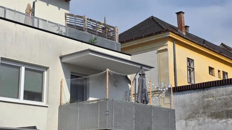 Moderne Wohnhausfassade mit einem Balkon und einer Dachterrasse, umgeben von älteren Gebäuden unter blauem Himmel.
