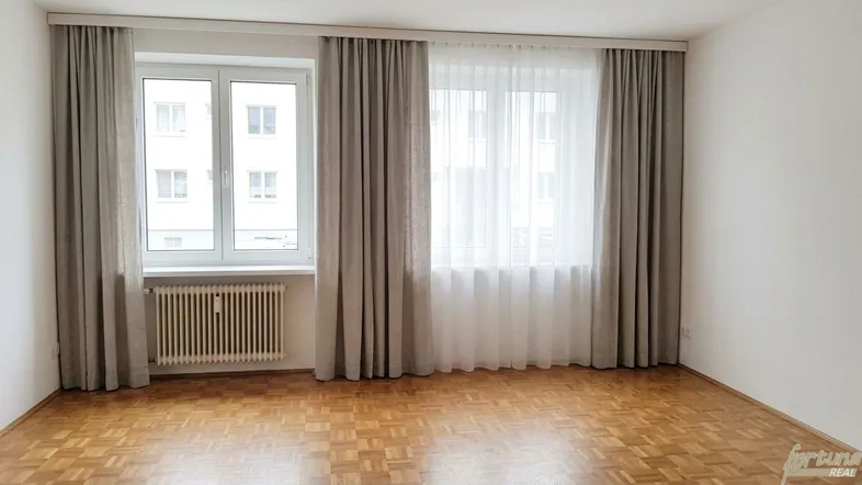 Helles Zimmer mit Parkettboden, weißen Wänden und großen Fenstern mit Vorhängen.
