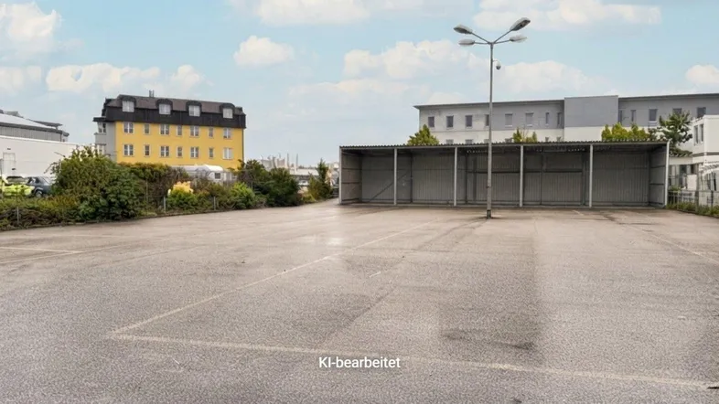 Großer asphaltierter Parkplatz mit einer offenen Überdachung und umliegenden Gebäuden unter bewölktem Himmel.
