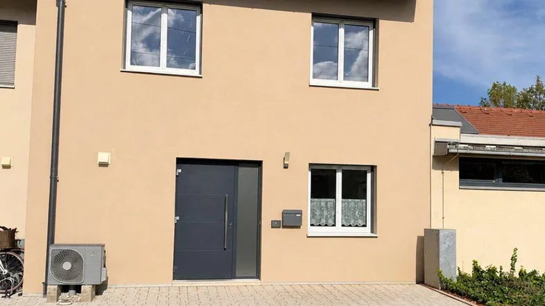 Moderne Hausfassade in hellem Beige mit dunkler Eingangstür und gepflastertem Vorplatz.
