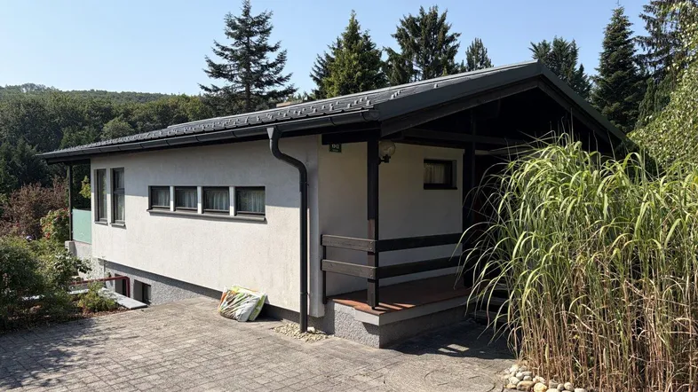 Hausfassade mit gepflasterter Auffahrt und umgebender Natur unter blauem Himmel.