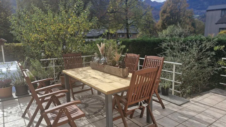 Geräumige Terrasse mit Holztisch und Stühlen, umgeben von üppigem Grün und Bergblick.