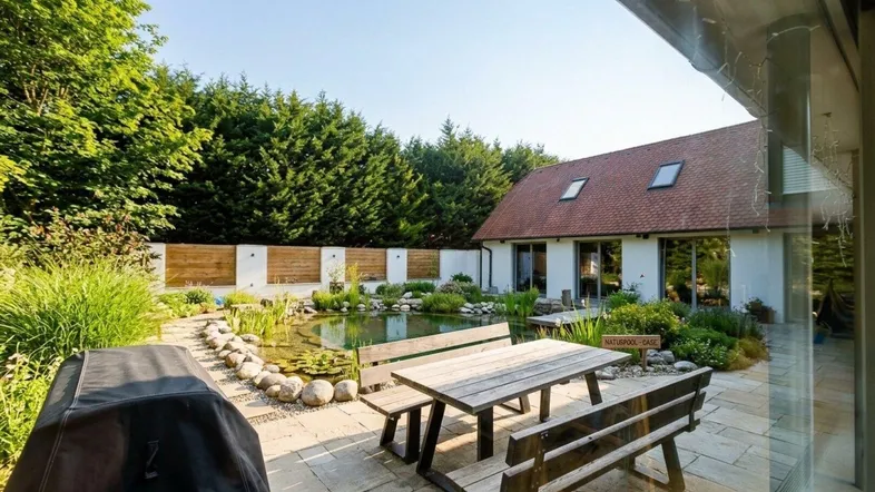 Große Terrasse mit Esstisch und Bänken, umgeben von einem Teich und üppigem Grün.