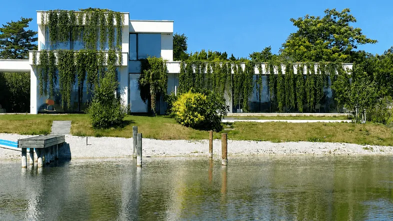 Moderne Villa am Seeufer mit üppiger Begrünung und privatem Steg unter blauem Himmel.