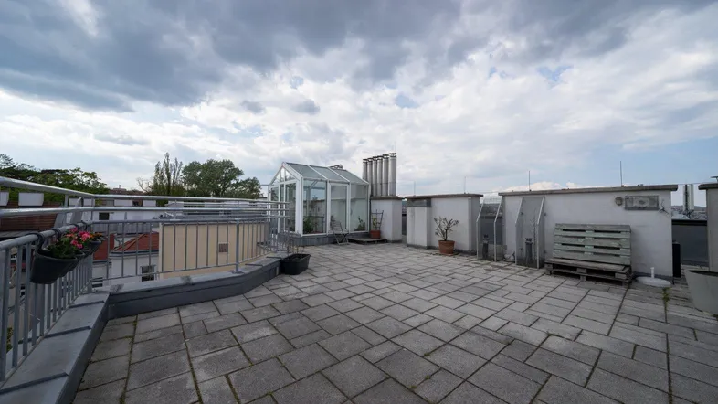 Geräumige Dachterrasse mit Steinplatten und einer kleinen Glasstruktur unter bewölktem Himmel.
