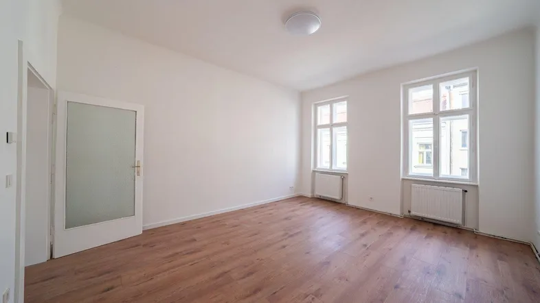 Geräumiges, helles Zimmer mit zwei Fenstern, Holzboden und weiß gestrichenen Wänden.