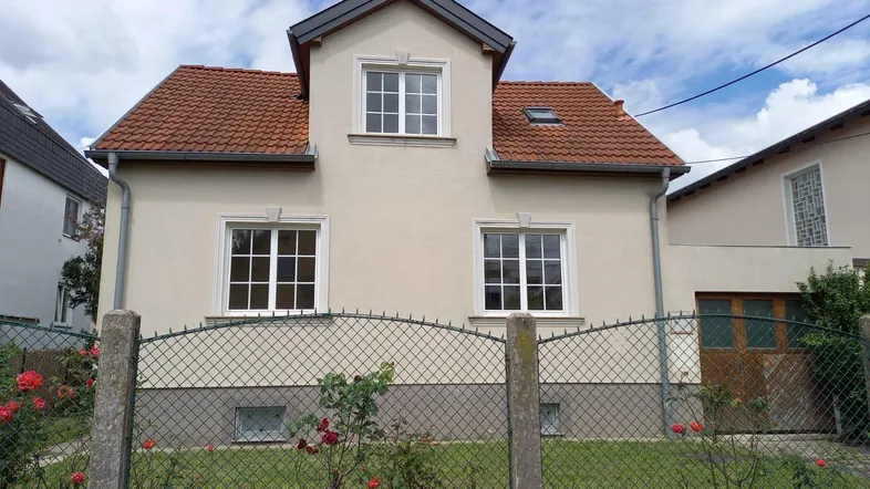 Vorderansicht des Hauses mit hellbeiger Fassade, roten Dachziegeln und einem kleinen Vorgarten.