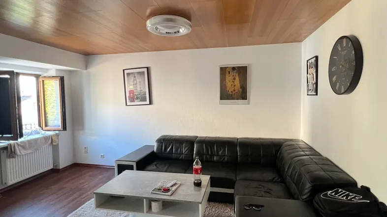 Geräumiges Wohnzimmer mit dunklem Laminatboden, Holzdecke und einer großen schwarzen Ledercouch.