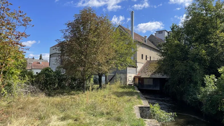 Altes Industriegebäude am Flussufer mit viel Grün und einem Schornstein unter blauem Himmel.