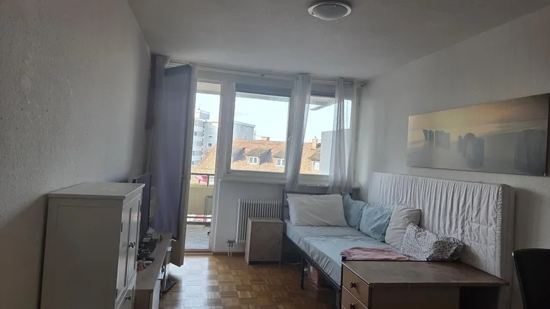Heller Wohn- und Schlafbereich mit Parkettboden, Schreibtisch und Zugang zu einem Balkon.
