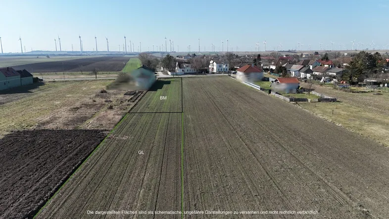Luftaufnahme eines ländlichen Gebiets mit Feldern, einem Dorf und Windkraftanlagen im Hintergrund.